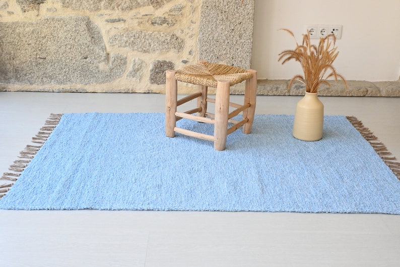 Pu&ograve; includere: Un tappeto blu chiaro con frange sui bordi. Uno sgabello di legno con una seduta intrecciata si trova sul tappeto. Un vaso con erba secca si trova sul tappeto.