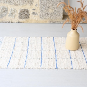 Pode incluir: Um tapete branco tecido com listras azuis. O tapete tem uma borda franjada e está sobre um piso cinza claro. Um vaso com grama seca está sobre o tapete.