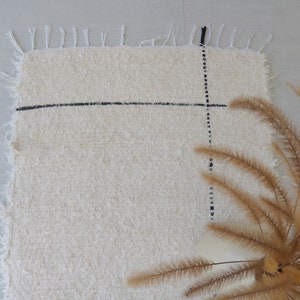 May include: A white rug with black lines forming a grid pattern. The rug has a fringe border and is lying on a light gray surface. A bouquet of dried brown grass is in the lower right corner of the image.
