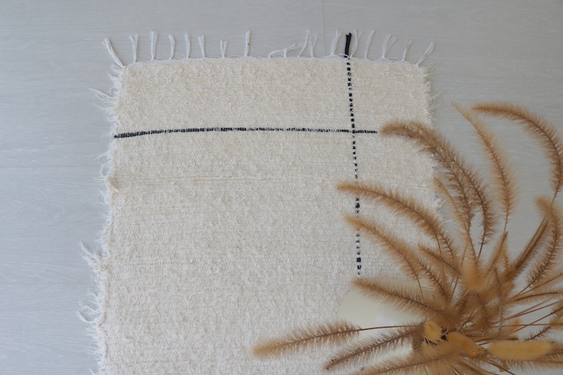 May include: A white rug with black lines forming a grid pattern. The rug has a fringe border and is lying on a light gray surface. A bouquet of dried brown grass is in the lower right corner of the image.