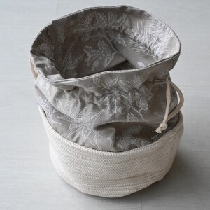 Pu&ograve; includere: Un cestino portaoggetti rotondo, aperto, con una base intrecciata bianco sporco e una parte superiore in tessuto grigio. Il tessuto grigio presenta un motivo floreale bianco e una chiusura con coulisse. Il cestino &egrave; progettato per lo stoccaggio.