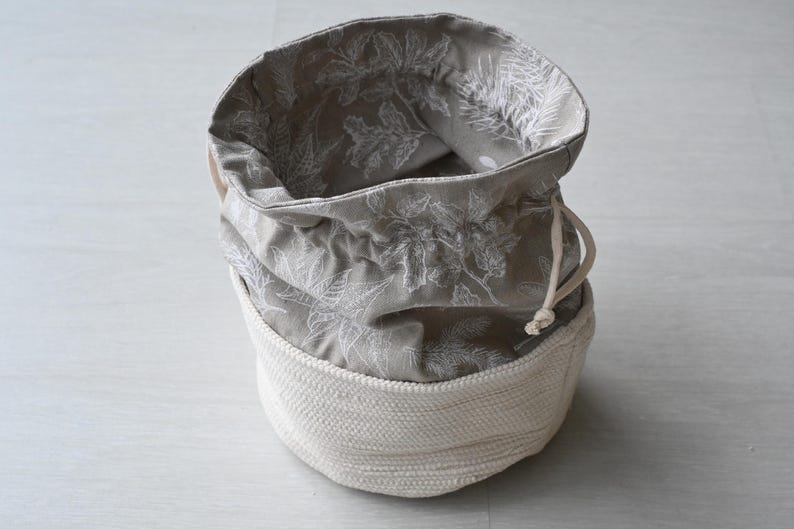 Pu&ograve; includere: Un cestino portaoggetti rotondo, aperto, con una base intrecciata bianco sporco e una parte superiore in tessuto grigio. Il tessuto grigio presenta un motivo floreale bianco e una chiusura con coulisse. Il cestino &egrave; progettato per lo stoccaggio.