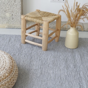 May include: A gray woven rug with fringe on the edges. A woven stool with a natural wood frame and a woven seat sits on the rug. A round woven cushion sits on the rug. A vase with dried grass sits on the rug.