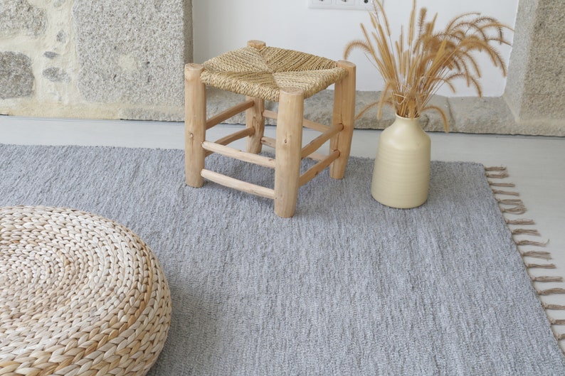 May include: A gray woven rug with fringe on the edges. A woven stool with a natural wood frame and a woven seat sits on the rug. A round woven cushion sits on the rug. A vase with dried grass sits on the rug.