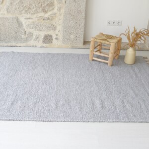 May include: A gray woven rug with fringe on all sides. The rug is laid out on a white floor with a small wooden stool and a vase with dried grass in the background.