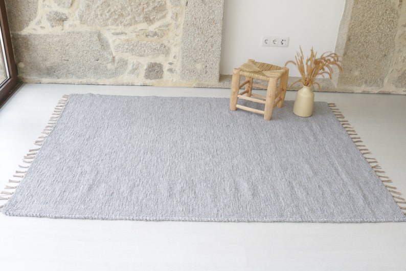 May include: A gray woven rug with fringe on all sides. The rug is laid out on a white floor with a small wooden stool and a vase with dried grass in the background.