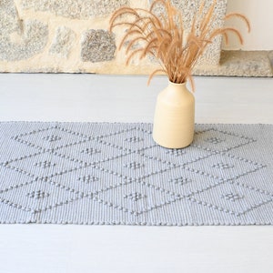Puede incluir: Una alfombra tejida gris con un patr&oacute;n de diamantes. La alfombra tiene una superficie texturizada y se coloca sobre un suelo blanco. Un jarr&oacute;n con hierba seca est&aacute; en el fondo.