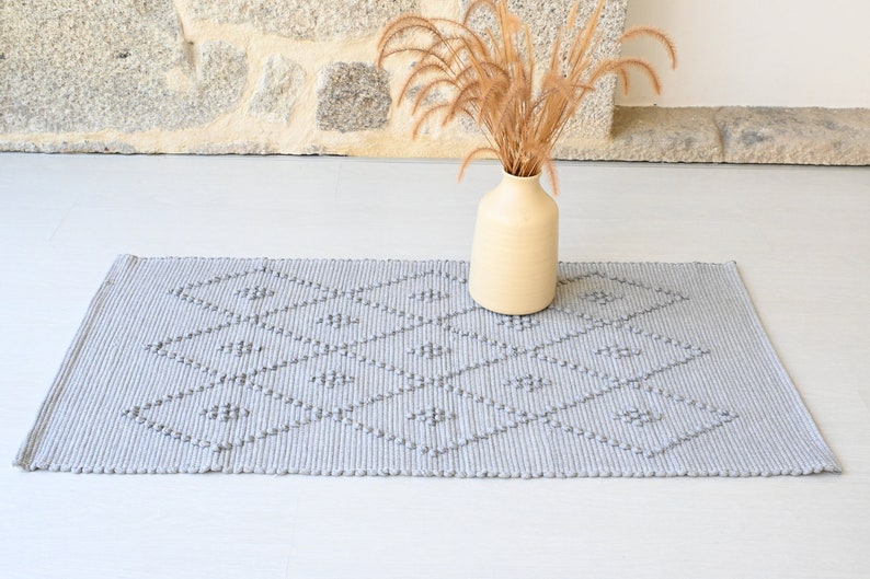 Puede incluir: Una alfombra tejida gris con un patr&oacute;n de diamantes. La alfombra tiene una superficie texturizada y se coloca sobre un suelo blanco. Un jarr&oacute;n con hierba seca est&aacute; en el fondo.