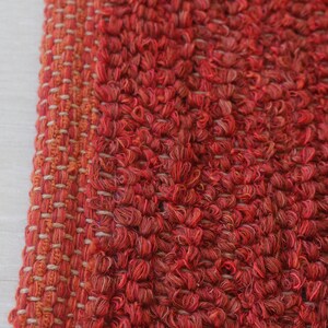 May include: Close-up of a red and orange woven rug with a textured surface. The rug has a fringe border.