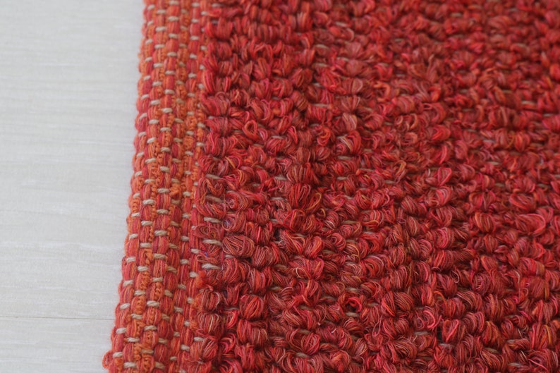 May include: Close-up of a red and orange woven rug with a textured surface. The rug has a fringe border.