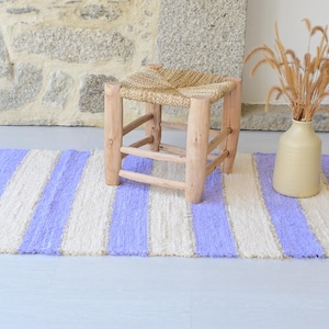 Könnte beinhalten: Ein gestreifter Teppich mit lavendelfarbenen und cremefarbenen Streifen. Ein kleiner Holzschemel mit geflochtener Sitzfläche steht darauf. Eine beige Vase mit getrockneten Gräsern ergänzt die Szene.