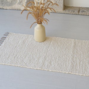 Puede incluir: Una alfombra blanca y esponjosa con flecos en los bordes. La alfombra est&aacute; colocada sobre un suelo gris claro con un jarr&oacute;n de hierba seca en el fondo.