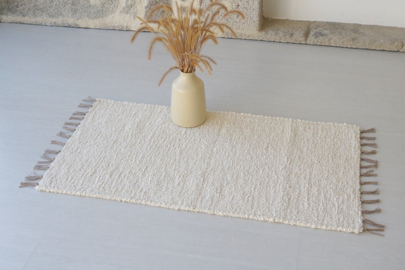 Puede incluir: Una alfombra blanca y esponjosa con flecos en los bordes. La alfombra est&aacute; colocada sobre un suelo gris claro con un jarr&oacute;n de hierba seca en el fondo.