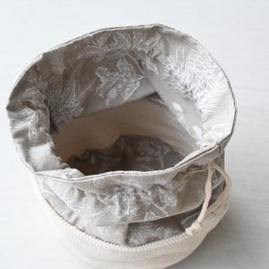 Pu&ograve; includere: Un piccolo cestino portaoggetti in tessuto aperto con chiusura a coulisse. L'interno &egrave; grigio chiaro con un motivo floreale bianco. L'esterno &egrave; in corda beige chiaro.