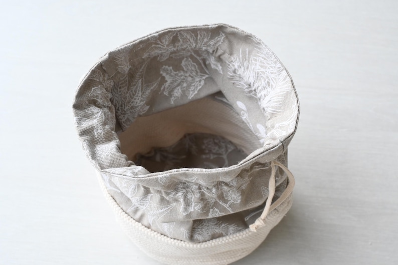 Pu&ograve; includere: Un piccolo cestino portaoggetti in tessuto aperto con chiusura a coulisse. L'interno &egrave; grigio chiaro con un motivo floreale bianco. L'esterno &egrave; in corda beige chiaro.