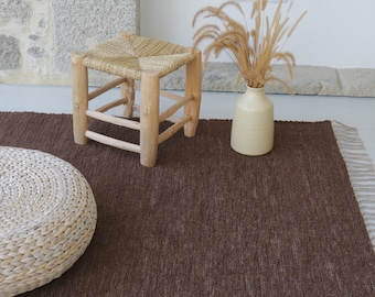 May include: A brown woven rug with fringe edges, a white woven stool, and a white woven pouf. The rug is on a light wood floor.