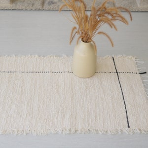 Op de afbeelding: Crèmekleurige geweven loper met een zwart rasterpatroon. Het tapijt heeft een gefranjerde rand en wordt gecombineerd met een beige vaas met gedroogde grassen. Een decoratief woonaccessoire.