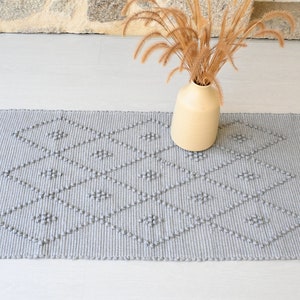 Puede incluir: Una alfombra tejida gris con un patr&oacute;n de diamantes y textura en relieve. La alfombra est&aacute; sobre un suelo gris claro con un jarr&oacute;n de hierba seca en el fondo.