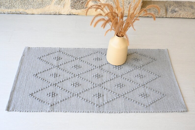 Puede incluir: Una alfombra tejida gris con un patr&oacute;n de diamantes y textura en relieve. La alfombra est&aacute; sobre un suelo gris claro con un jarr&oacute;n de hierba seca en el fondo.