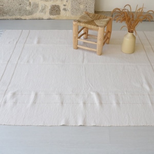 Puede incluir: Una alfombra blanca tejida con flecos en los bordes y rayas sutiles. La alfombra est&aacute; colocada sobre un suelo gris claro con un taburete de madera y un jarr&oacute;n de flores secas en el fondo.