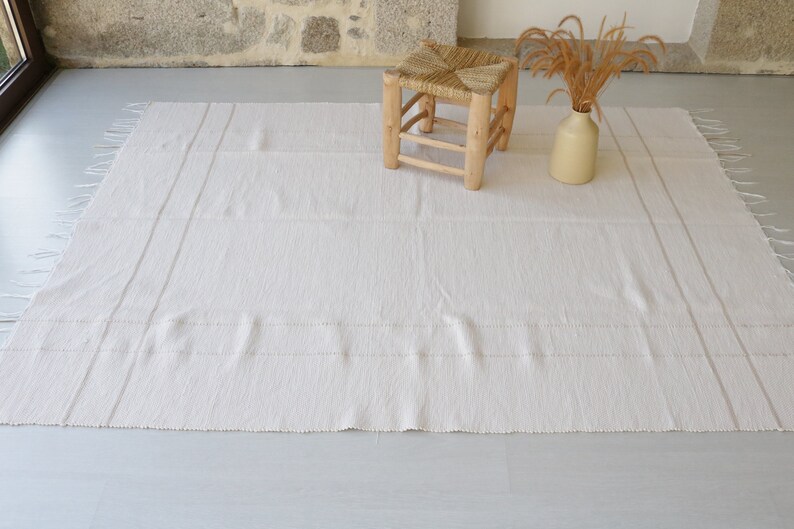 Puede incluir: Una alfombra blanca tejida con flecos en los bordes y rayas sutiles. La alfombra est&aacute; colocada sobre un suelo gris claro con un taburete de madera y un jarr&oacute;n de flores secas en el fondo.