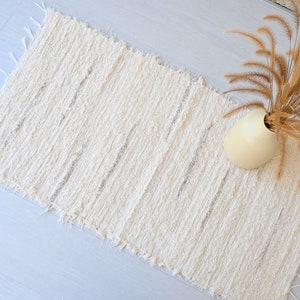May include: A white woven rug with a fringed edge. The rug has a subtle texture and is laid out on a light gray floor. A vase with dried grass is placed on the rug.