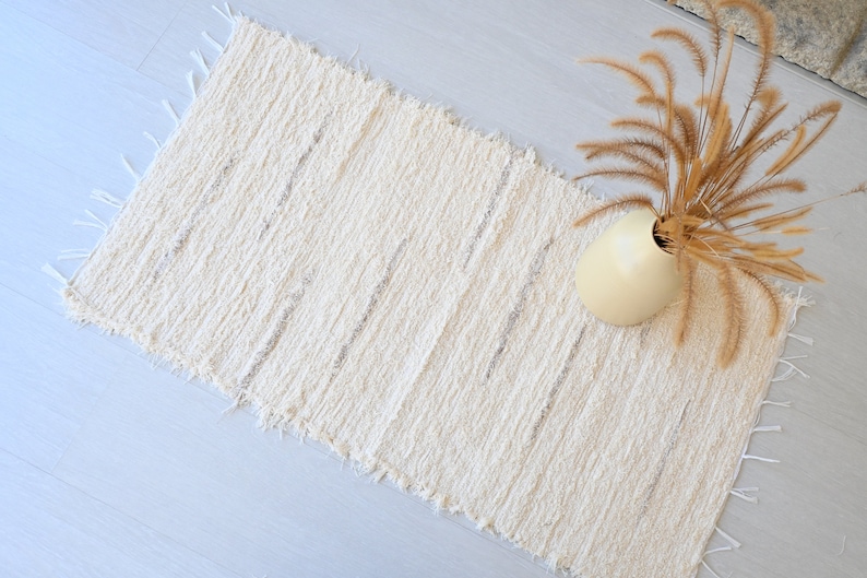 May include: A white woven rug with a fringed edge. The rug has a subtle texture and is laid out on a light gray floor. A vase with dried grass is placed on the rug.