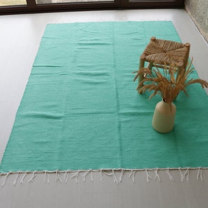 K&ouml;nnte beinhalten: Ein t&uuml;rkisfarbener gewebter Teppich mit wei&szlig;en Fransen. Ein kleiner Holzhocker mit einem gewebten Sitz und eine Vase mit getrocknetem Gras stehen auf dem Teppich.