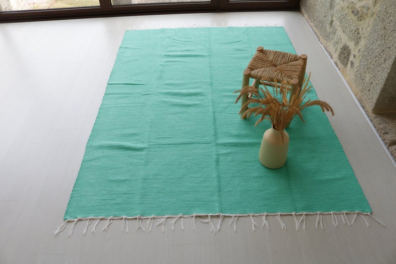 K&ouml;nnte beinhalten: Ein t&uuml;rkisfarbener gewebter Teppich mit wei&szlig;en Fransen. Ein kleiner Holzhocker mit einem gewebten Sitz und eine Vase mit getrocknetem Gras stehen auf dem Teppich.