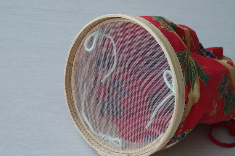 Pode incluir: Um peneira redonda de madeira com um forro de malha branca e la&ccedil;os de corda branca. A peneira &eacute; coberta com um tecido vermelho com um padr&atilde;o floral.