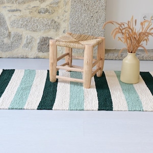 Peut inclure: Un tapis rayé avec des bandes alternées blanches, turquoise et vert foncé. Un petit tabouret en bois clair avec une assise tissée est posé sur le tapis. Un vase beige contient des plantes séchées.