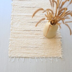 May include: A white woven rug with a fringe border. A vase with dried grass is on the rug.