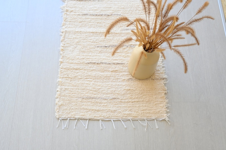 May include: A white woven rug with a fringe border. A vase with dried grass is on the rug.