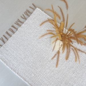 May include: White woven rug with fringe on the edges. The rug has a subtle texture and is shown with a vase of dried grass.