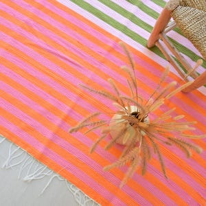 Op de afbeelding: Een rechthoekig geweven textiel met horizontale strepen in levendige tinten oranje, roze en groen, afgewerkt met witte franjes langs de rand. Een ronde vaas met gedroogde siergrassen staat op de stof bij een gevlochten krukje.