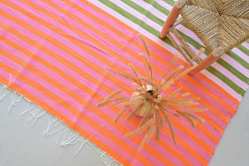 Op de afbeelding: Een rechthoekig geweven textiel met horizontale strepen in levendige tinten oranje, roze en groen, afgewerkt met witte franjes langs de rand. Een ronde vaas met gedroogde siergrassen staat op de stof bij een gevlochten krukje.