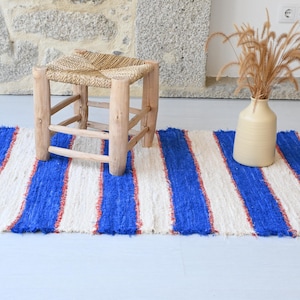 Può includere: Un tappeto rettangolare a righe blu e bianche, accentuato da sottili linee rosse. Un piccolo sgabello in legno con seduta intrecciata è posto sul tappeto. Un vaso beige contiene piante essiccate.