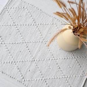 Pode incluir: Um tapete branco tecido com um padrão de diamantes. O tapete é feito de fio grosso e texturizado e tem um brilho sutil. Um vaso com grama seca é colocado sobre o tapete.