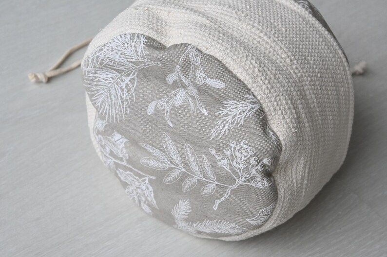 Pu&ograve; includere: Una borsa rotonda in tessuto bianco sporco con chiusura a coulisse. L'interno &egrave; grigio con una stampa botanica bianca di foglie e bacche. L'esterno &egrave; un materiale tessuto e strutturato.