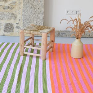 Op de afbeelding: Een kleine houten kruk met een gevlochten zitting, een beige keramische vaas met gedroogde grassen en een rechthoekig kleed met verticale strepen in groen, lichtpaars, feloranje en roze met witte franjes aan de randen.