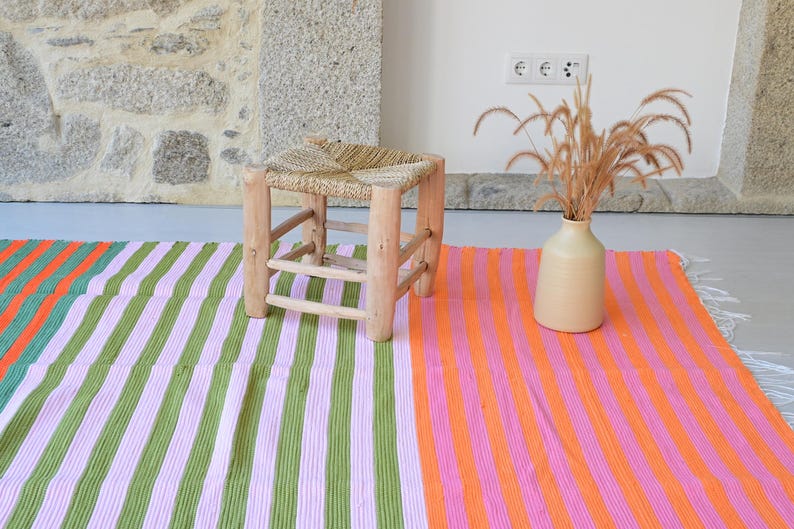 Op de afbeelding: Een kleine houten kruk met een gevlochten zitting, een beige keramische vaas met gedroogde grassen en een rechthoekig kleed met verticale strepen in groen, lichtpaars, feloranje en roze met witte franjes aan de randen.
