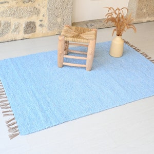 Pu&ograve; includere: Un tappeto blu chiaro con frange sui bordi. Il tappeto &egrave; realizzato in un materiale morbido e soffice e ha una texture sottile. Uno sgabello in legno con una seduta intrecciata si trova sopra il tappeto.