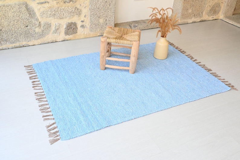 Pu&ograve; includere: Un tappeto blu chiaro con frange sui bordi. Il tappeto &egrave; realizzato in un materiale morbido e soffice e ha una texture sottile. Uno sgabello in legno con una seduta intrecciata si trova sopra il tappeto.
