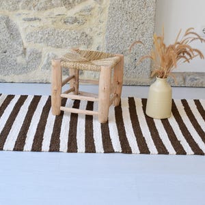 Può includere: Un tappeto rettangolare a righe marroni e bianche. Un piccolo sgabello in legno con seduta intrecciata è appoggiato sul tappeto. Un vaso beige con piante secche è a destra dello sgabello. Lo sfondo è un muro di pietra.