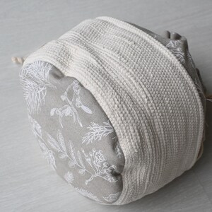 Pu&ograve; includere: Piccolo sacchetto rotondo in tessuto con chiusura a coulisse. Il tessuto grigio presenta un motivo botanico bianco. Una spessa fascia intrecciata bianco sporco avvolge il centro. Il cordino &egrave; beige naturale.