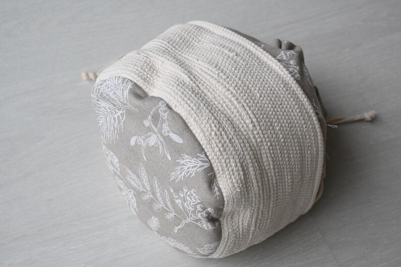 Pu&ograve; includere: Piccolo sacchetto rotondo in tessuto con chiusura a coulisse. Il tessuto grigio presenta un motivo botanico bianco. Una spessa fascia intrecciata bianco sporco avvolge il centro. Il cordino &egrave; beige naturale.