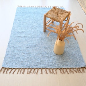 Pu&ograve; includere: Un tappeto tessuto di colore azzurro chiaro con frange sui bordi. Il tappeto &egrave; su un pavimento in legno chiaro e ha uno sgabello di legno con una seduta intrecciata e un vaso con erbe secche sopra.