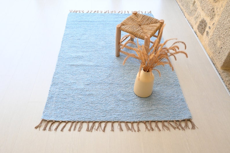 Pu&ograve; includere: Un tappeto tessuto di colore azzurro chiaro con frange sui bordi. Il tappeto &egrave; su un pavimento in legno chiaro e ha uno sgabello di legno con una seduta intrecciata e un vaso con erbe secche sopra.