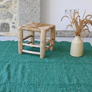 May include: A green woven rug with white fringe, a wooden stool with a woven seat, and a vase with dried grass.