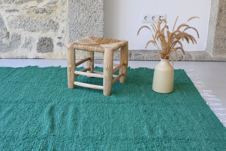 May include: A green woven rug with white fringe, a wooden stool with a woven seat, and a vase with dried grass.